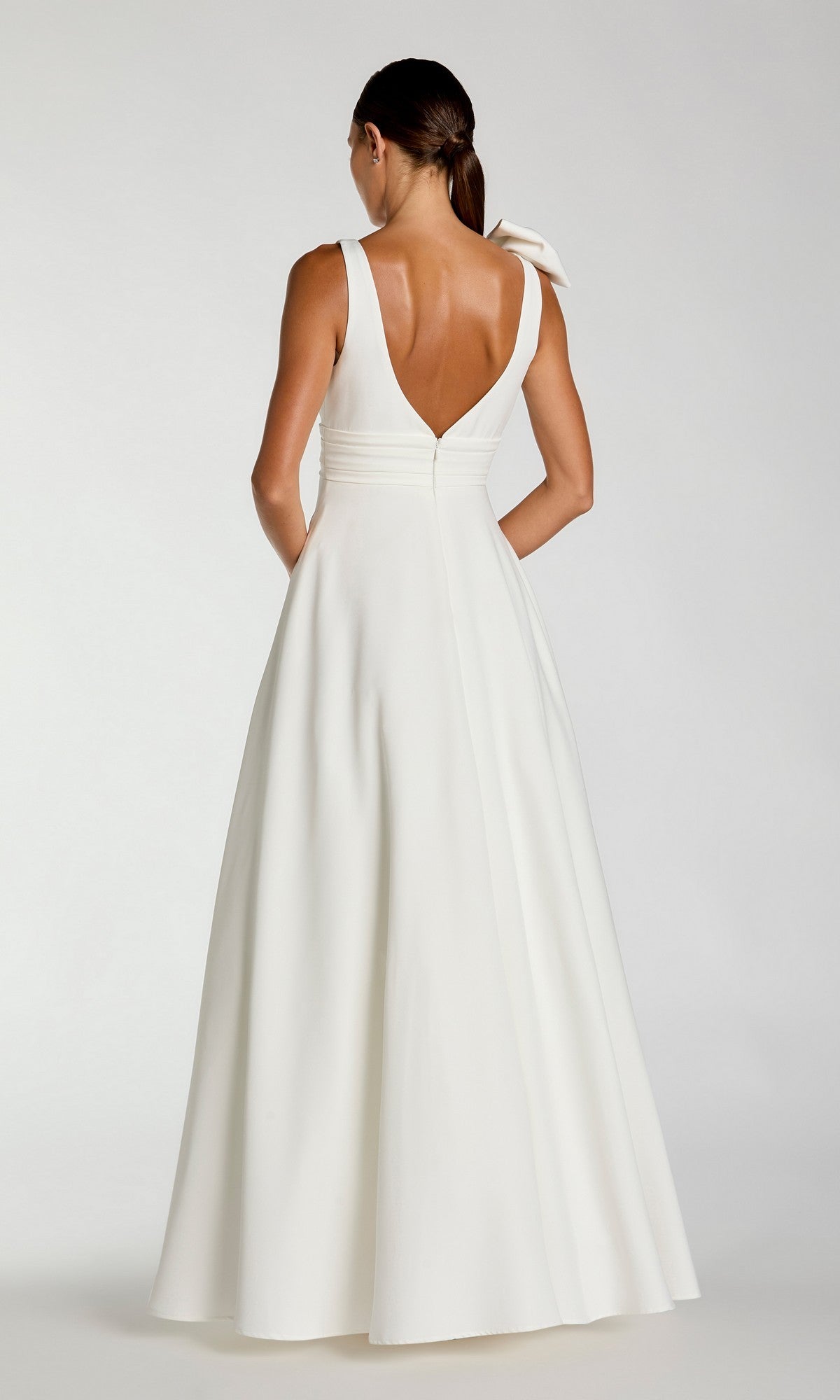 A woman in the Mac Duggal 49972 Shoulder-Bow Long Formal Dress, a sleeveless white gown with a V-back and full skirt, stands facing away with her hair tied back in a low ponytail.